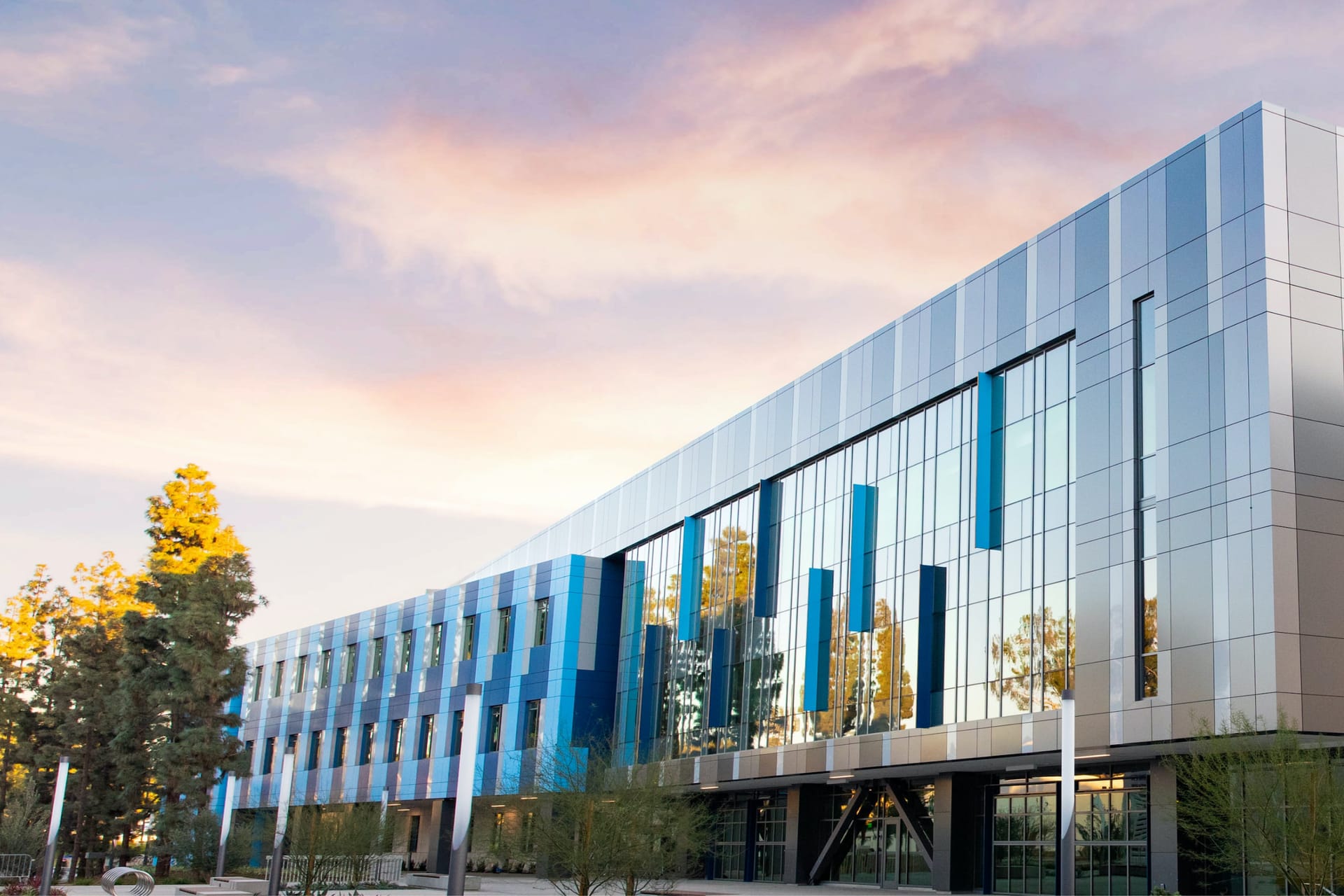 Modern glass office building with blue panels reflecting sunset sky, surrounded by trees with autumn foliage.