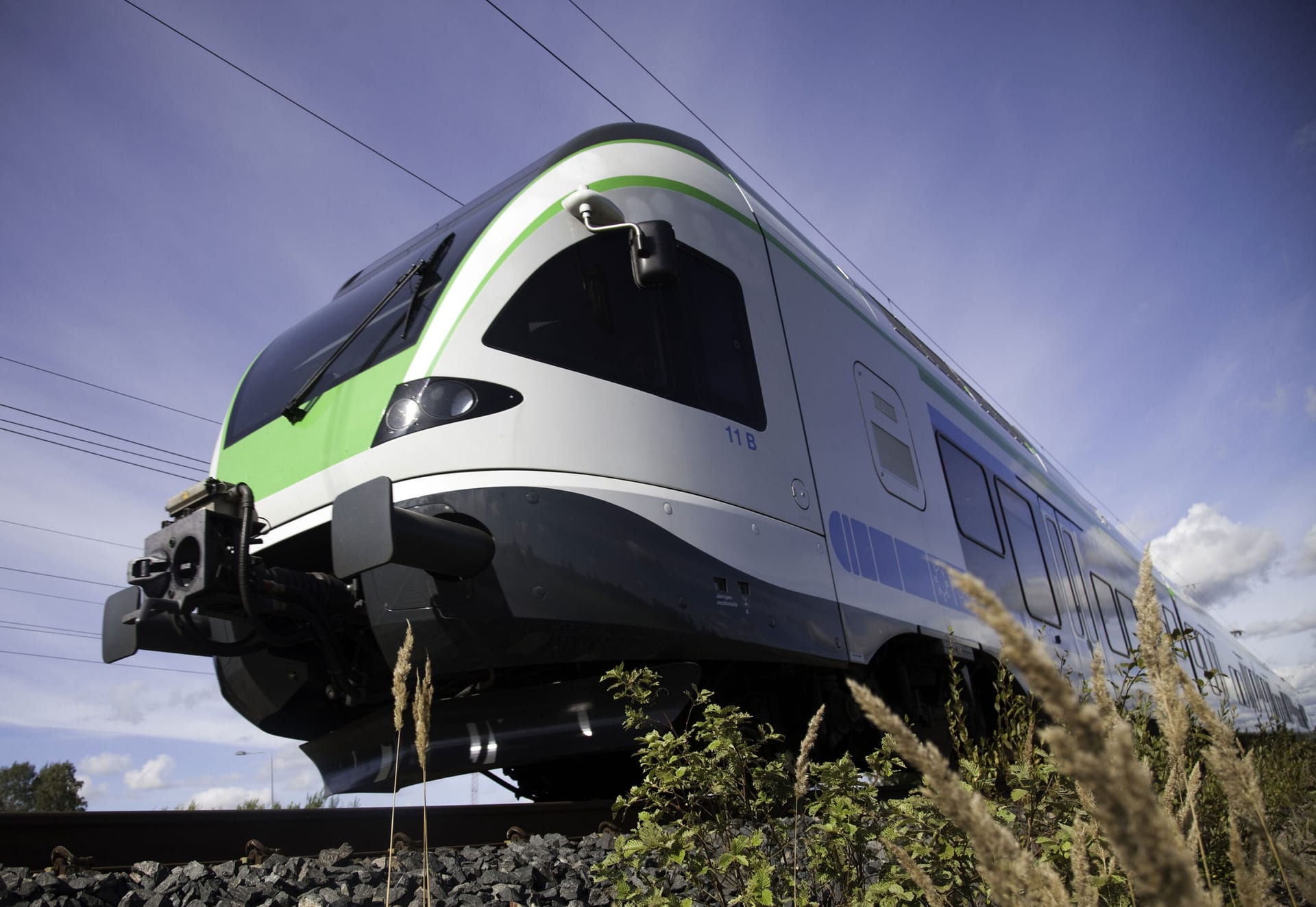 Modern passenger train with white and green livery travelling on tracks against blue sky, viewed from low angle.