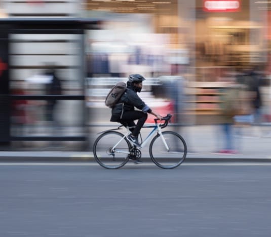 Cyclist with backpack riding a blue bicycle through a city street, captured with motion blur effect.