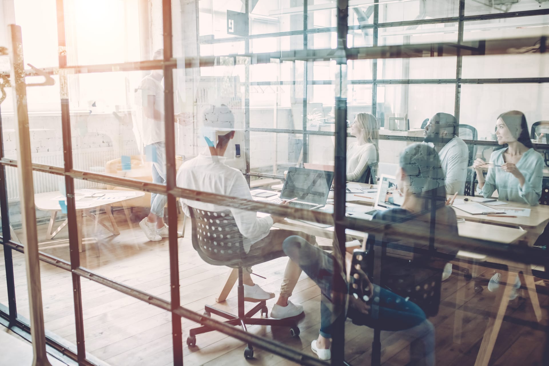 Modern office meeting viewed through glass partition, with diverse professionals collaborating around laptops in sunlit space.