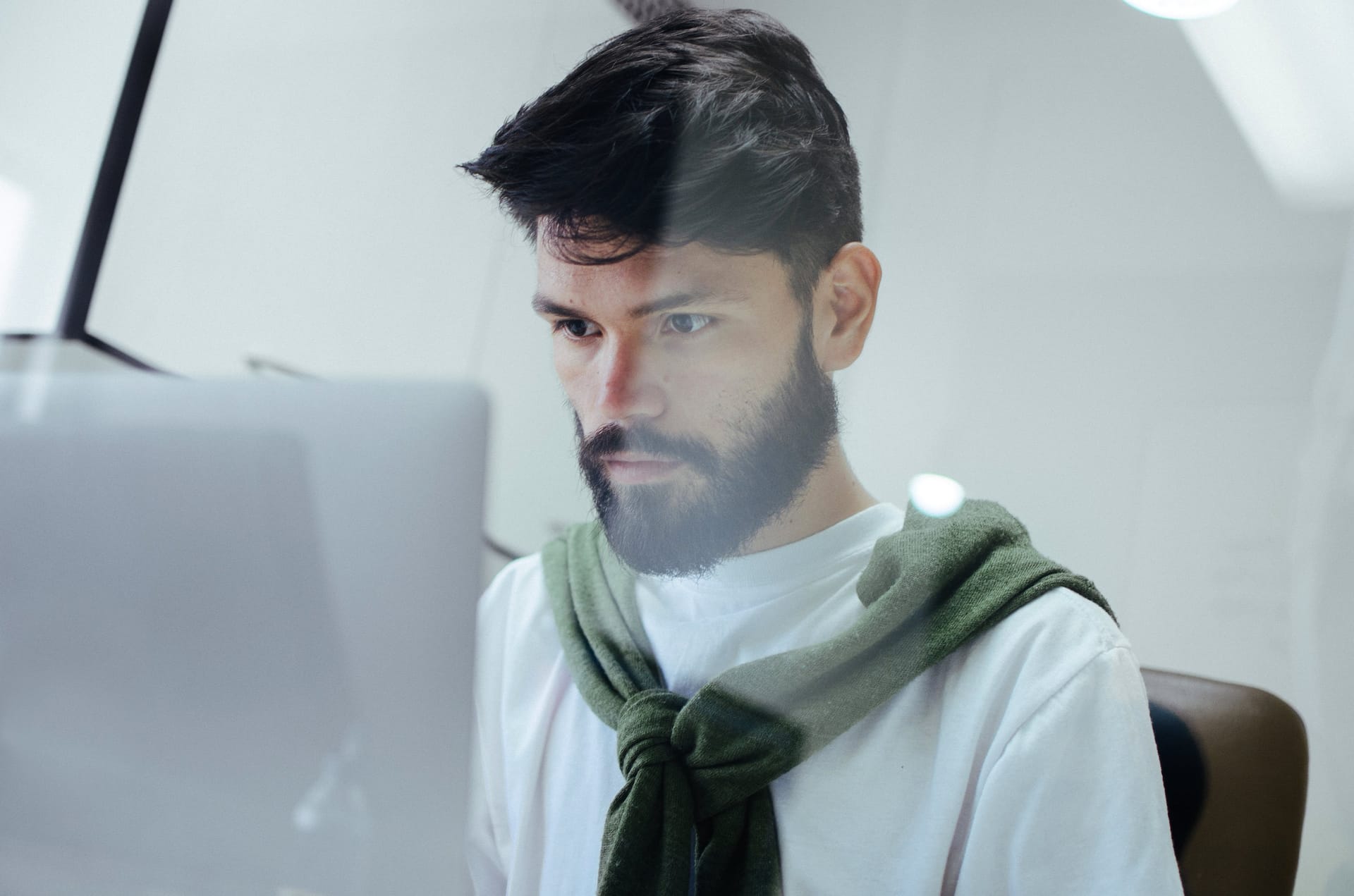 Person with dark hair and beard wearing white shirt with green sweater tied around shoulders, focused on computer screen.