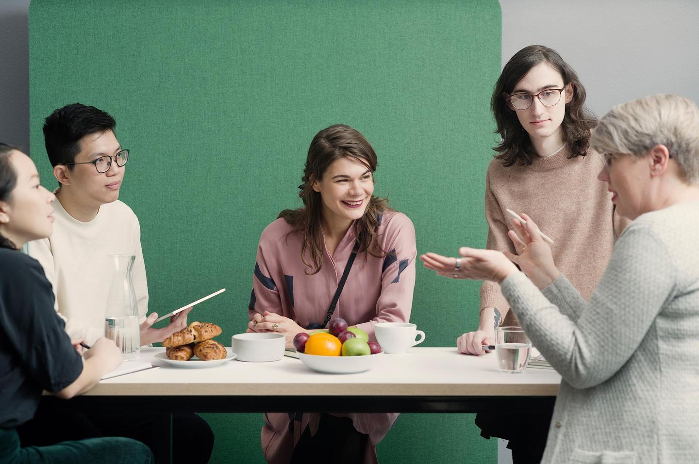 A diverse group of five is having an engaging conversation in the office. They are standing at a table in front of a green wall.