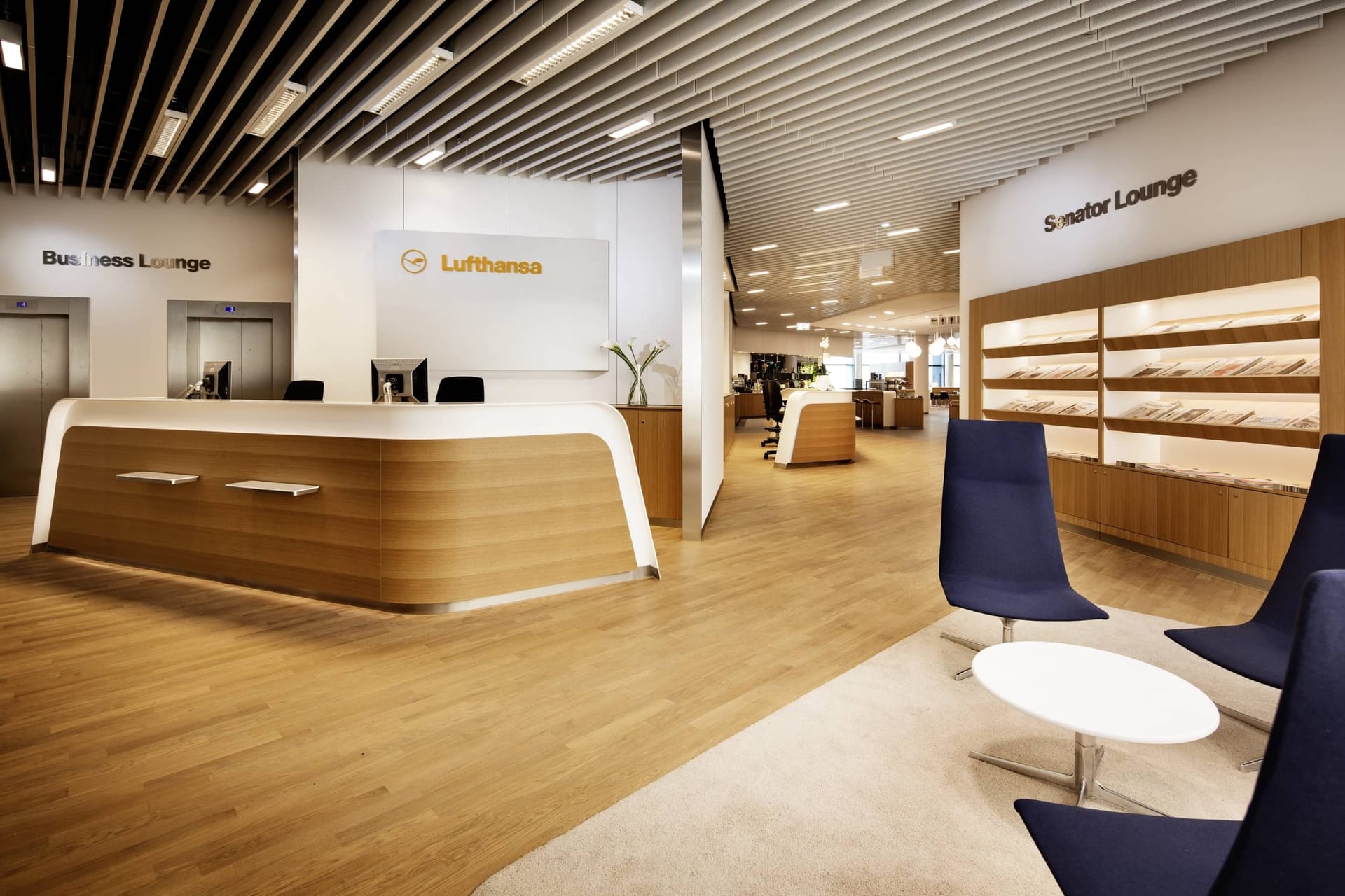 Modern Lufthansa airport lounge with wooden reception desk, blue chairs, and separate Business and Senator lounge areas.