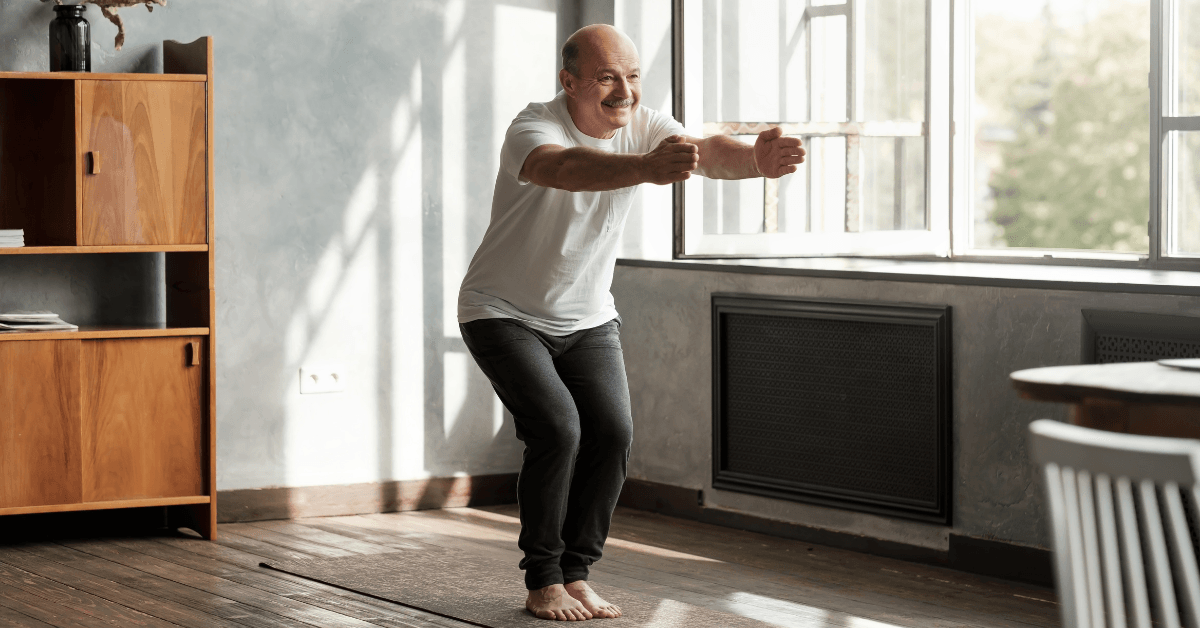 Older man in white t-shirt exercising in a bright room with wooden floors and furniture, smiling while stretching.
