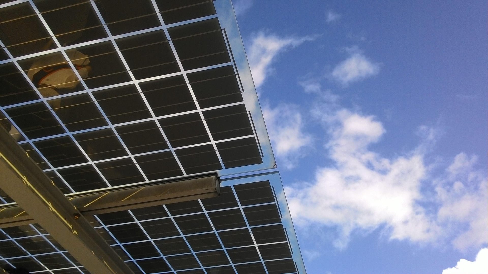 Solar panels mounted on a metal frame against a blue sky with scattered white clouds, viewed from below.