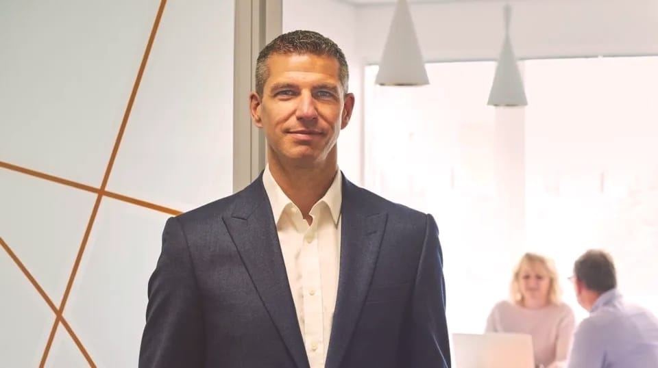 Professional in navy suit and white shirt standing in modern office with pendant lights and colleagues in background.