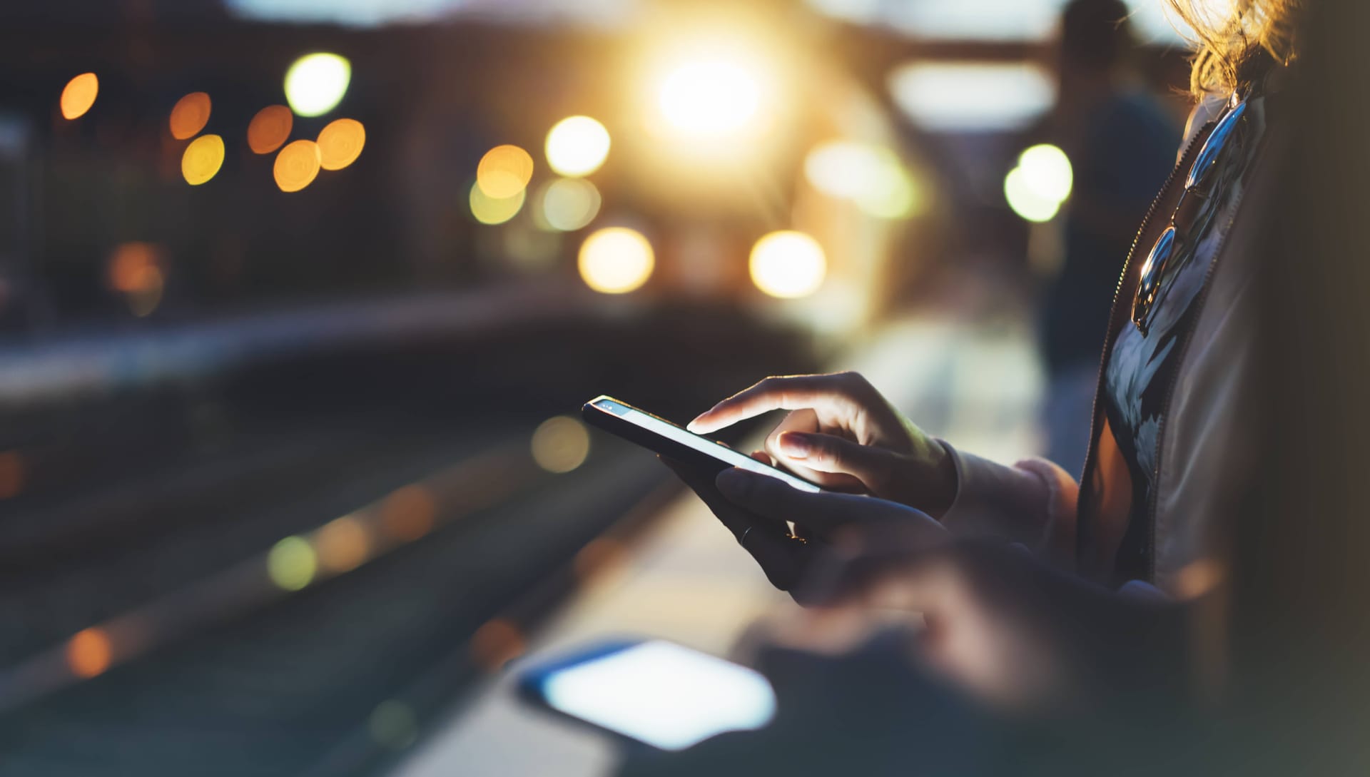 Person using smartphone at night with blurred city lights in background, creating atmospheric urban scene.