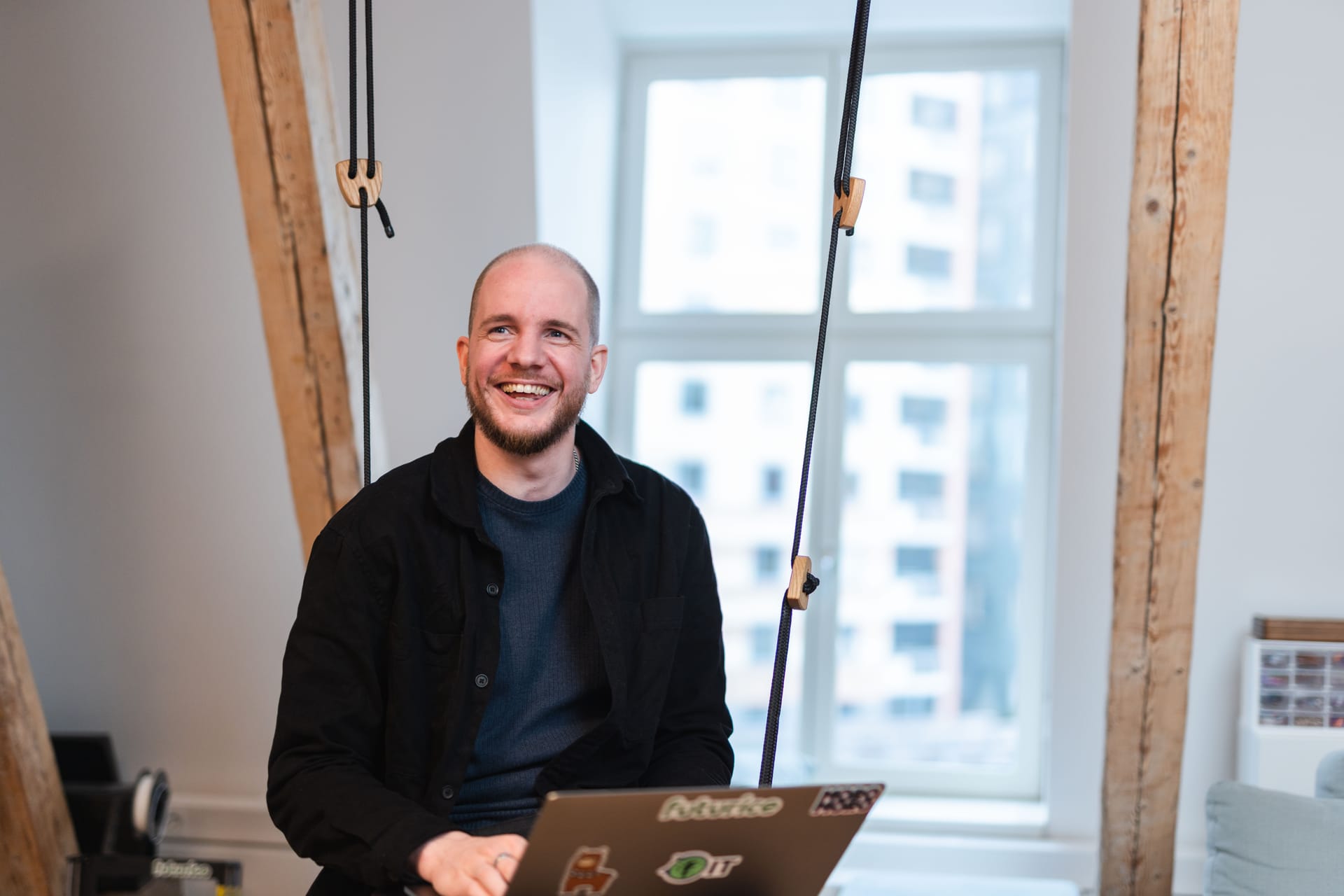 Person smiling in bright modern office with wooden beams, laptop open, large windows showing city buildings in background.