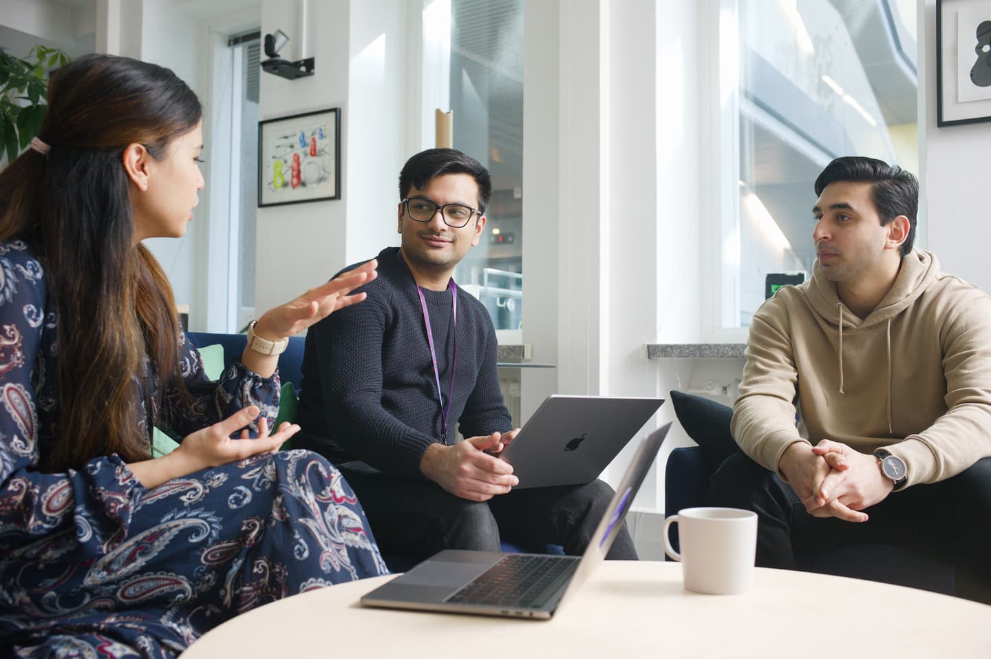 Three people in a bright office meeting space discussing ideas with laptops and coffee on the table.