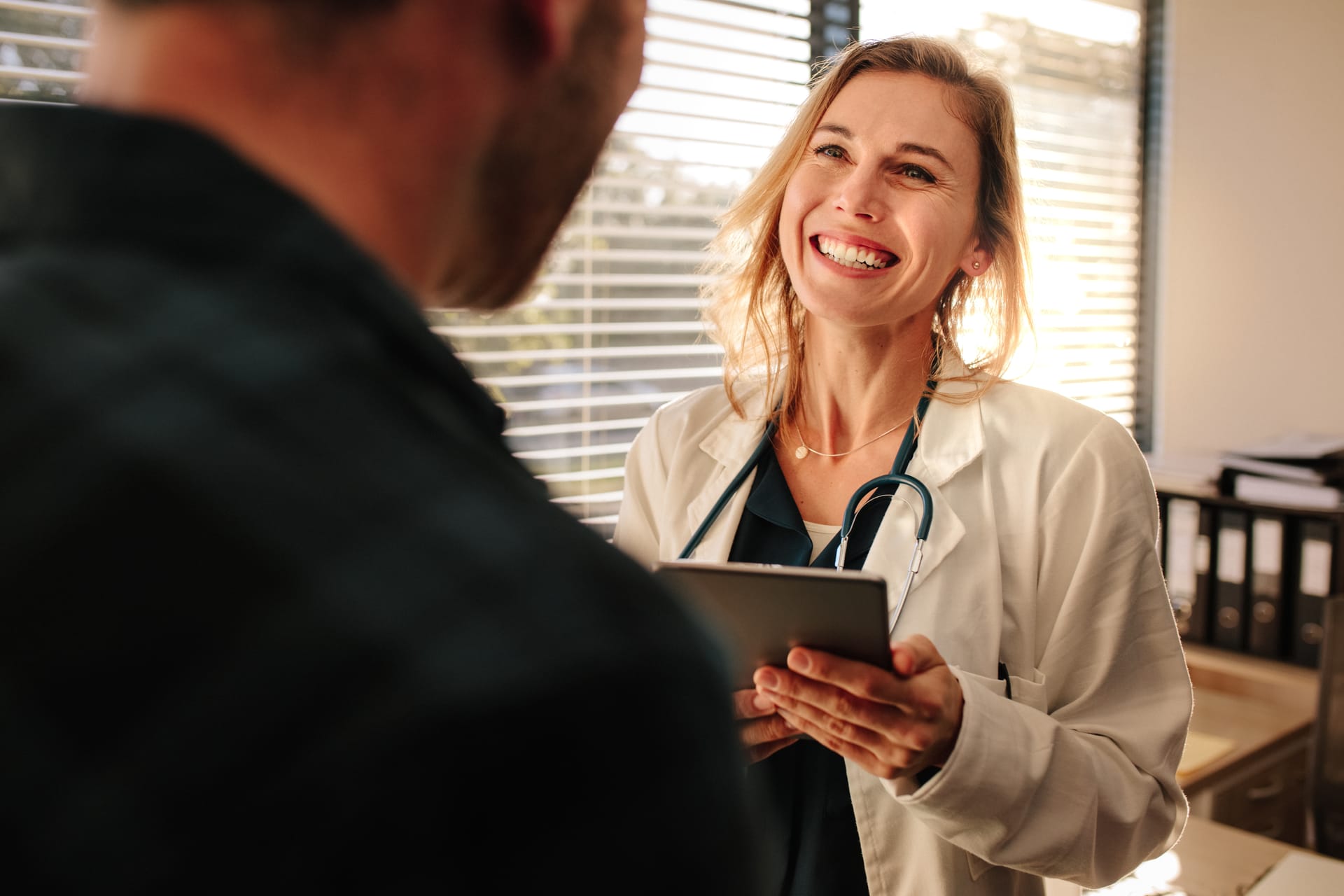 Smiling doctor in white coat with stethoscope holding tablet while speaking with patient in sunlit medical office.