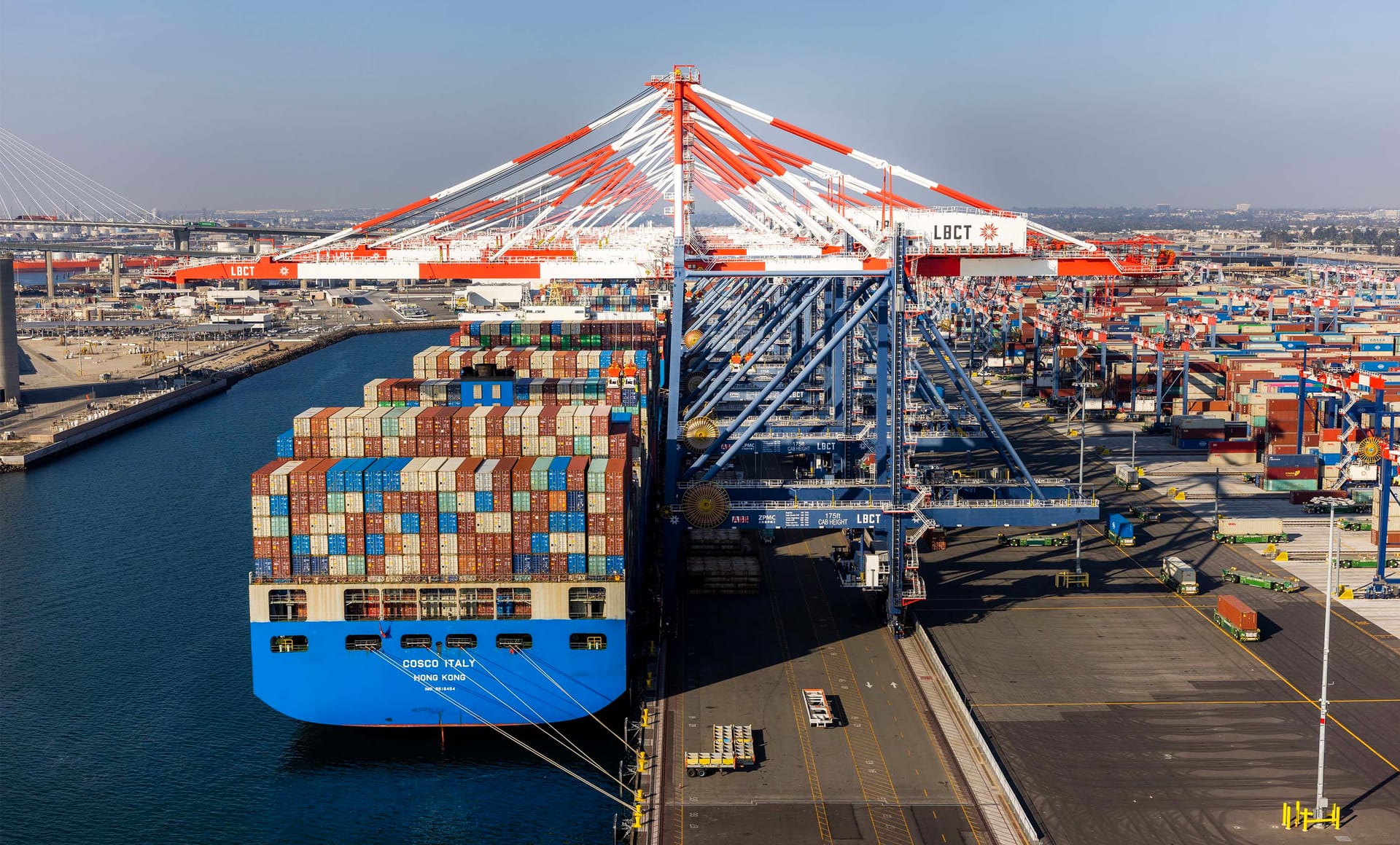 Large container ship docked at LBCT port terminal with stacked shipping containers and distinctive orange cranes overhead.