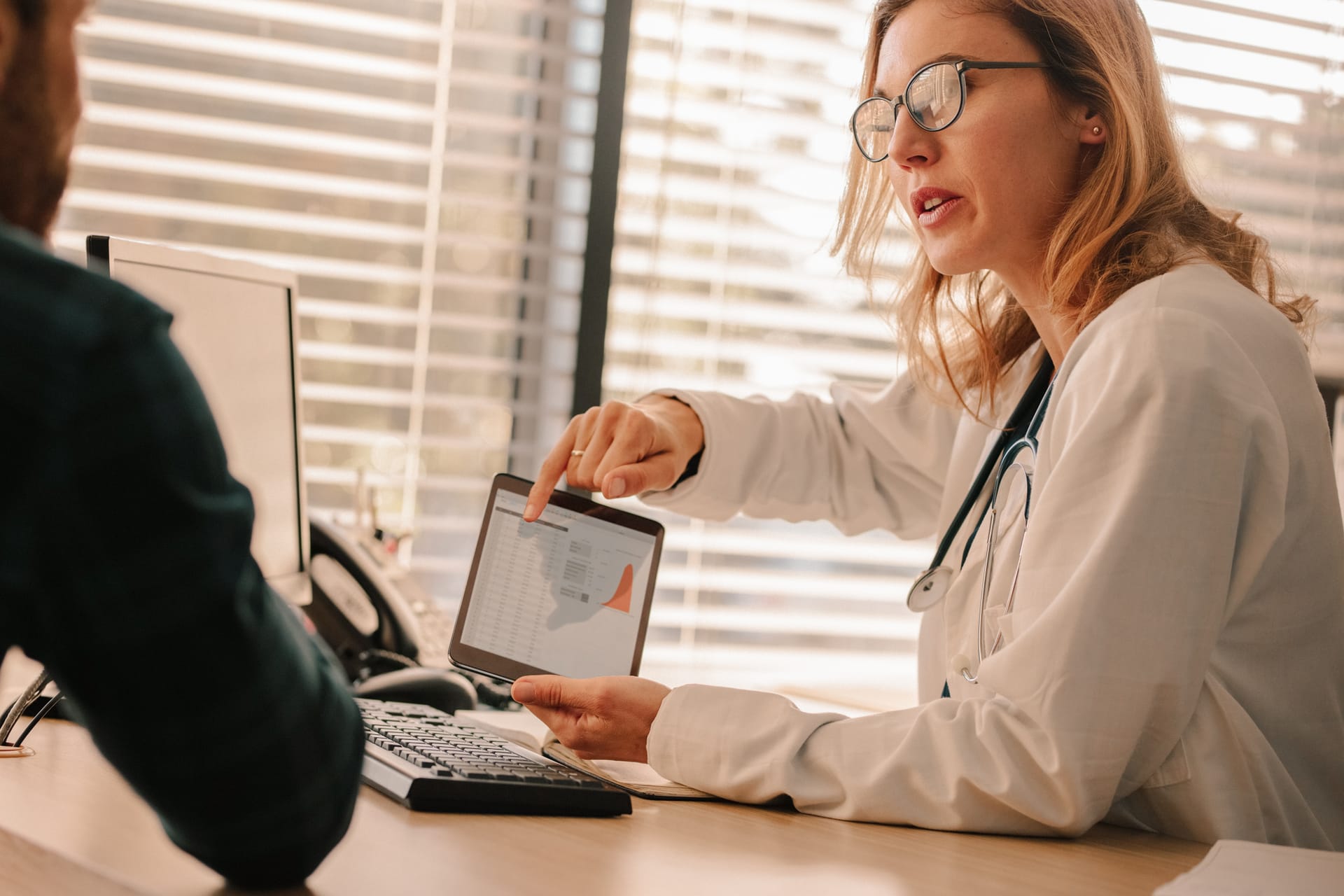 Doctor with stethoscope explaining medical data on tablet to patient during consultation near window with blinds.