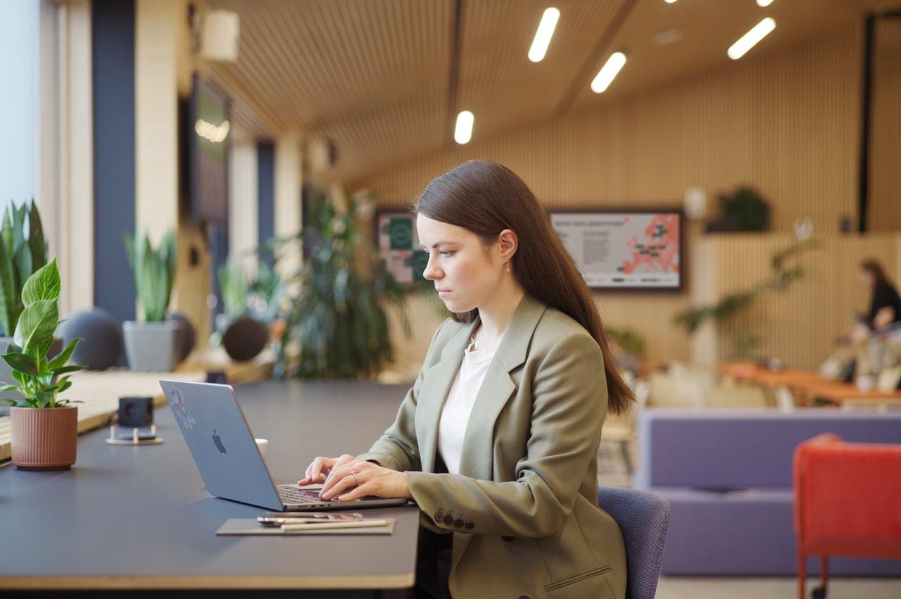 Person in green blazer working on laptop in modern office space with plants and warm lighting.