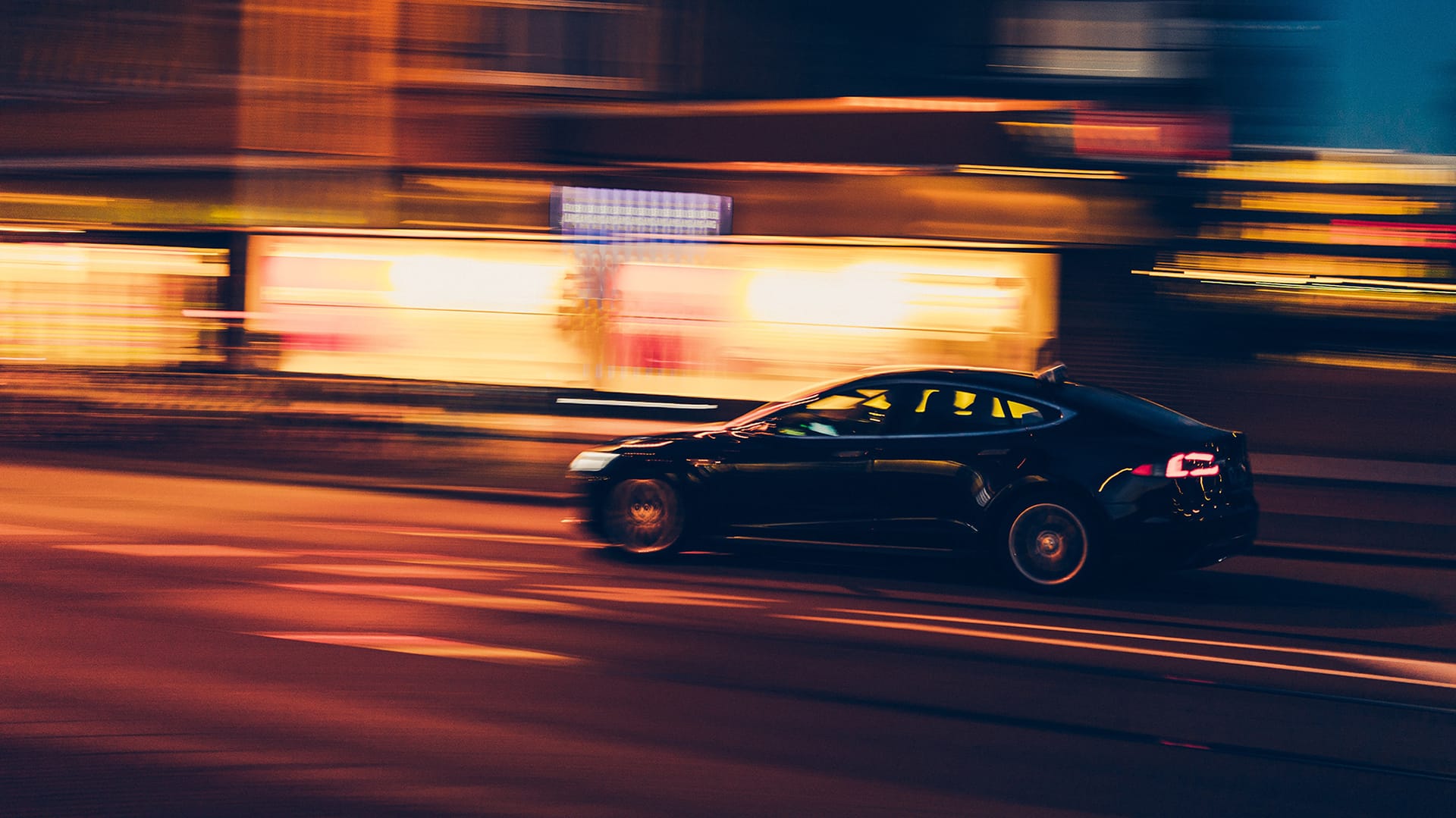 Black car speeding through city streets at night with motion blur and illuminated storefronts in the background.