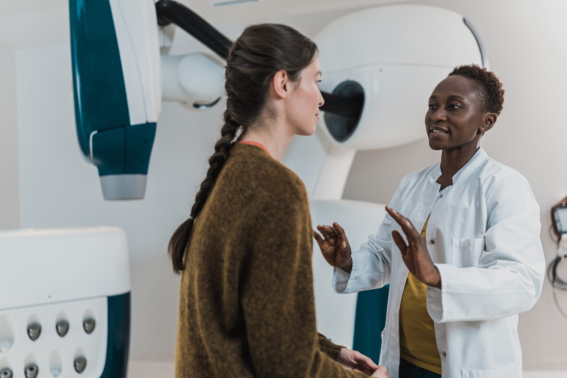 Healthcare professional in white coat explaining procedure to patient in brown sweater beside medical imaging equipment.