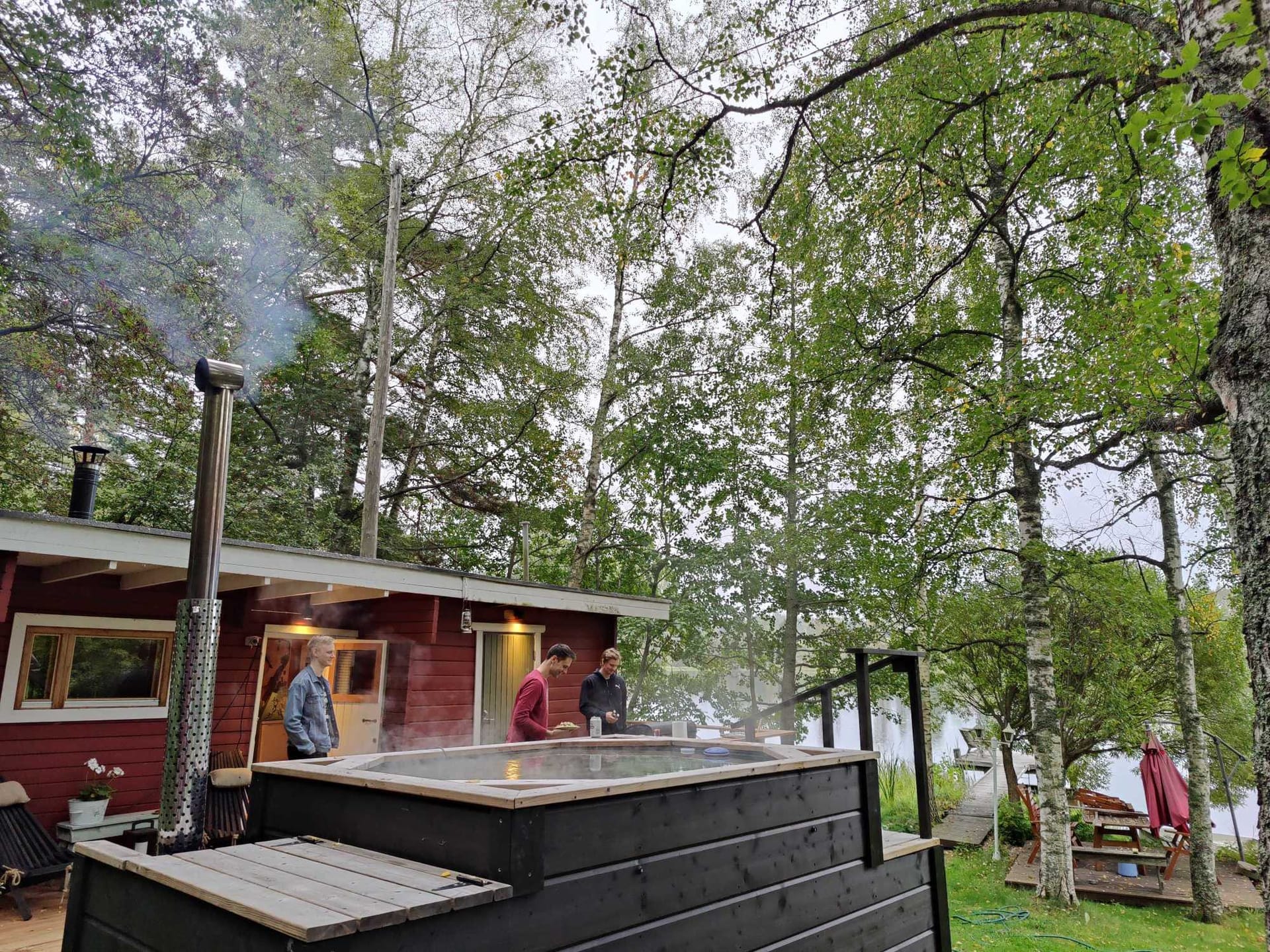 People enjoying an outdoor hot tub beside a red lakeside cabin surrounded by birch trees in a forest setting.
