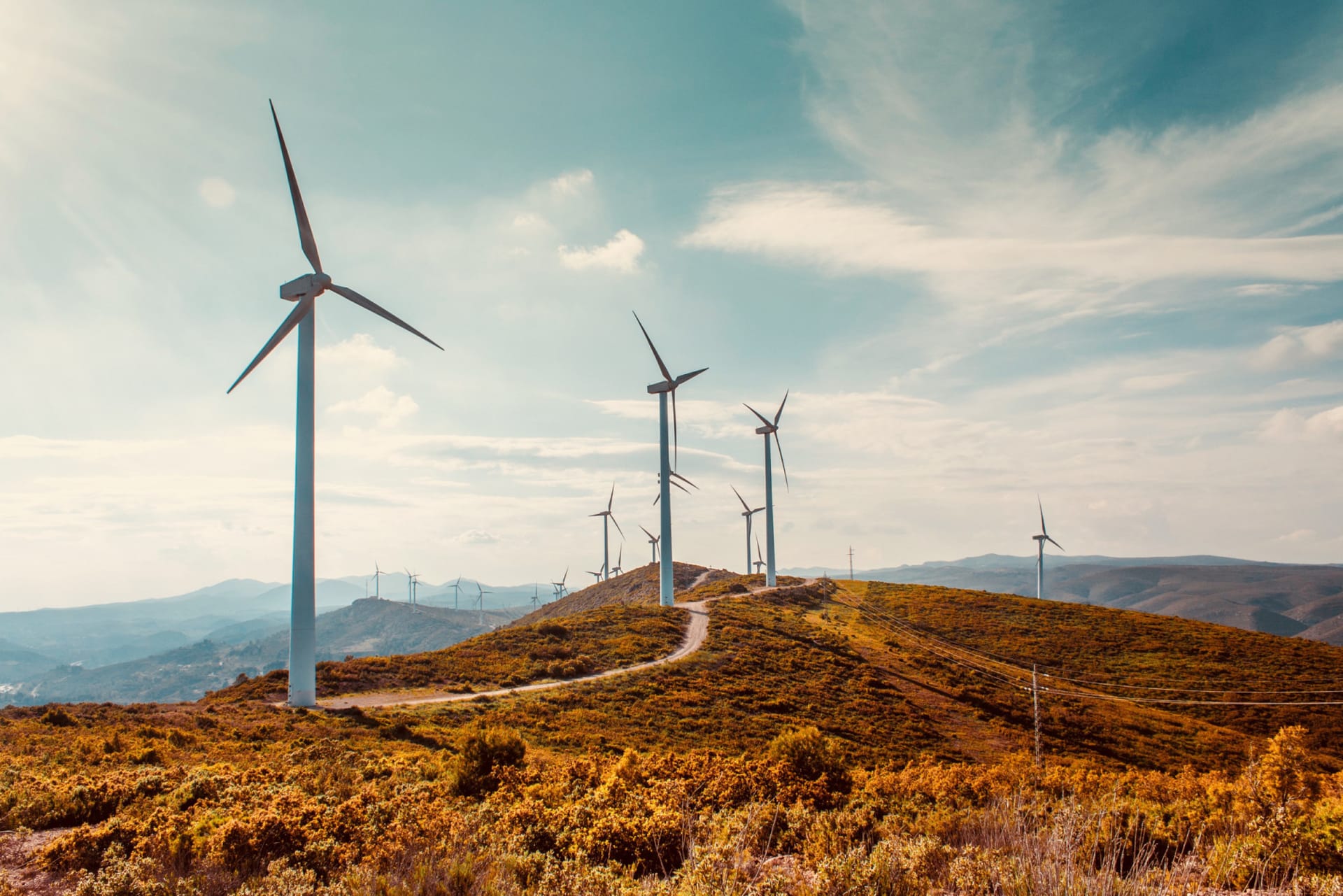 Wind turbines on golden hillside under blue sky, with winding path and mountain ranges in the background.
