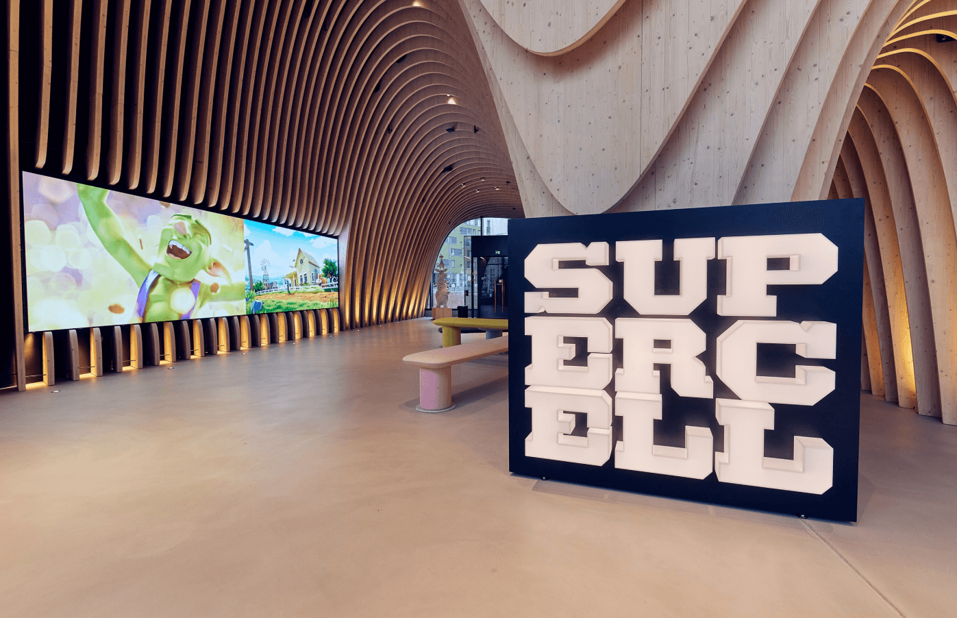 Modern exhibition space with curved wooden walls, large video screen showing animated character, and SUPERCELL sign.