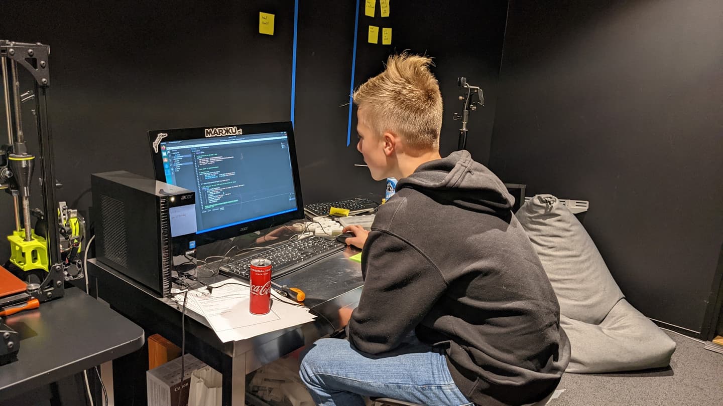 Person in grey hoodie coding on computer with Coca-Cola can nearby in dark workspace with yellow sticky notes on wall.