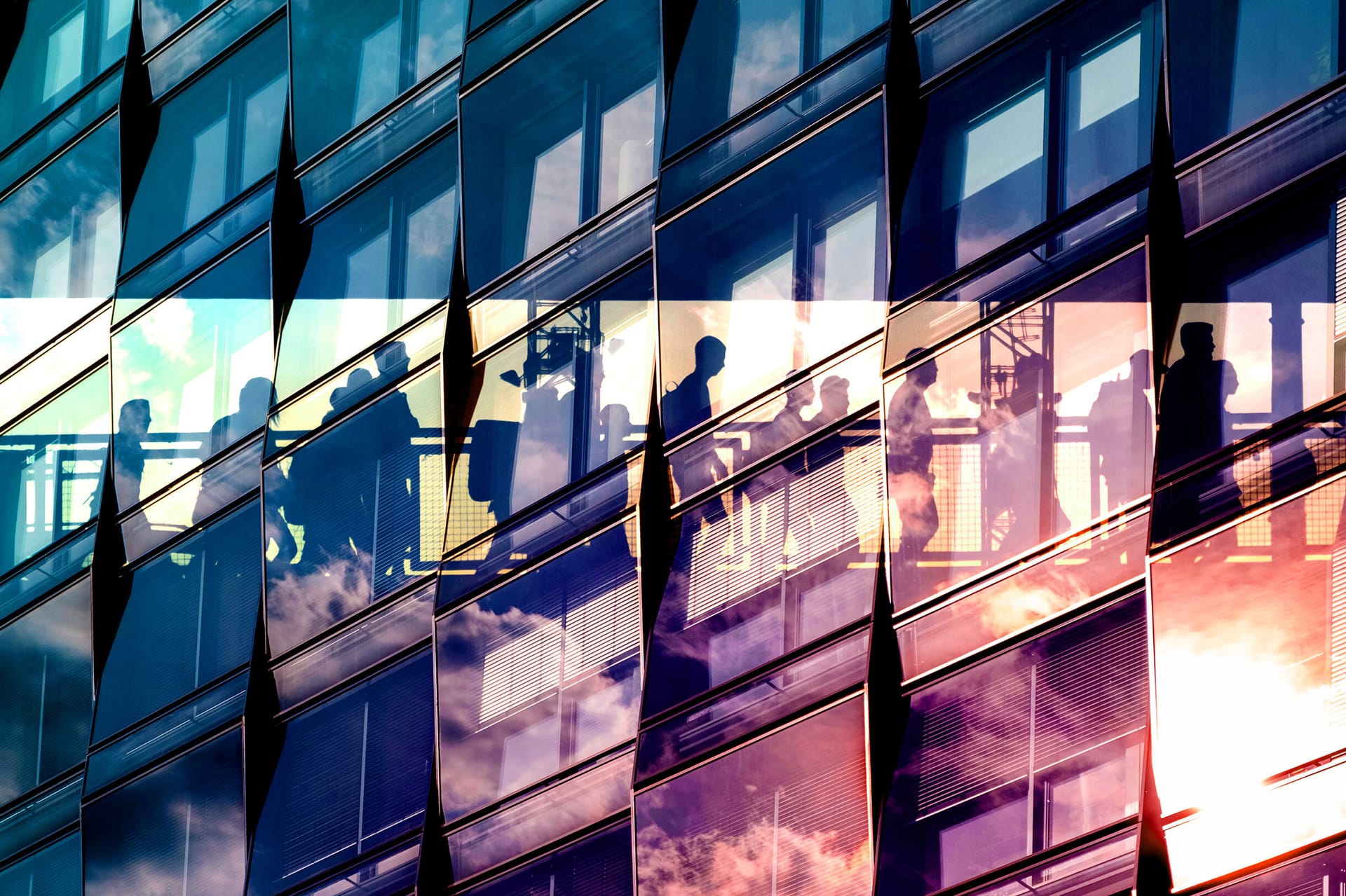 Silhouettes of people visible through modern glass building facade with blue and purple reflections of sky and clouds.