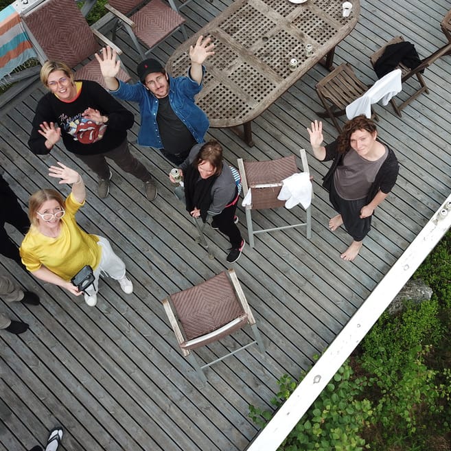 Aerial view of people waving from a wooden deck with outdoor furniture, surrounded by greenery.