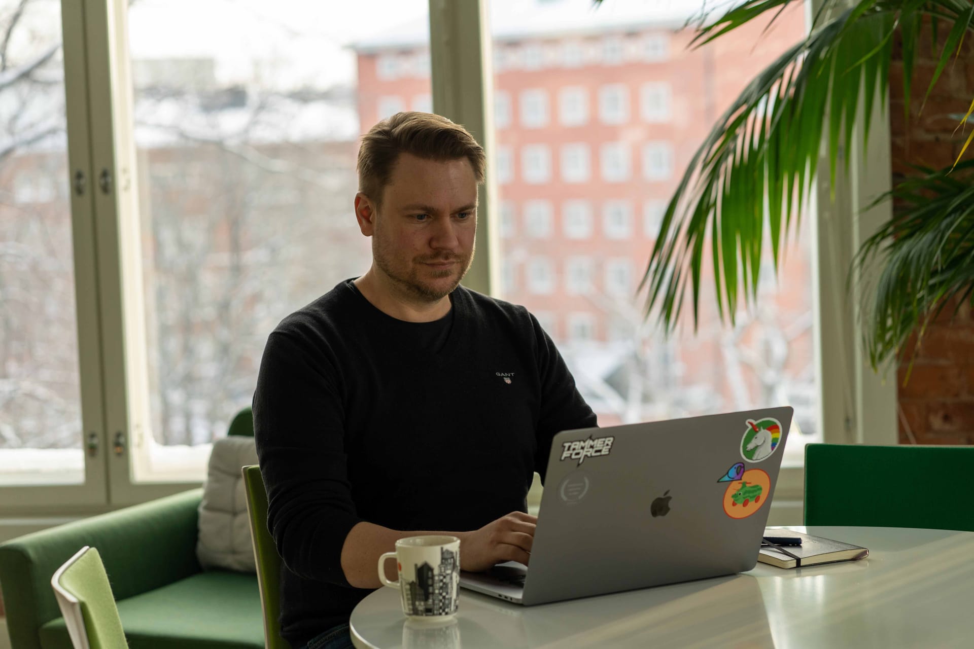 Person in black jumper working on laptop with stickered cover, coffee mug nearby, bright window view of brick building.
