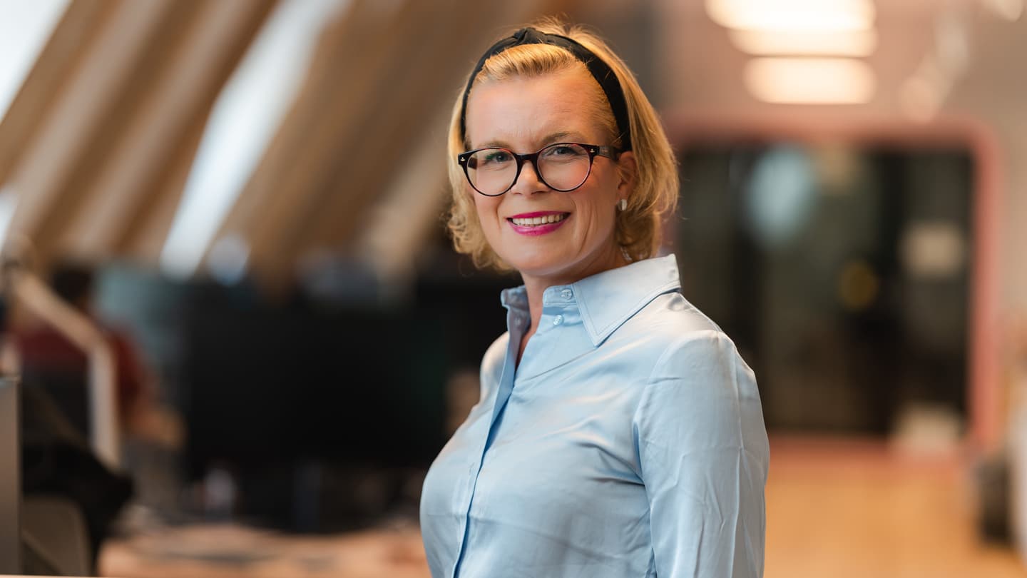 Professional woman with blonde hair wearing glasses, black headband and light blue shirt, smiling in an office setting.