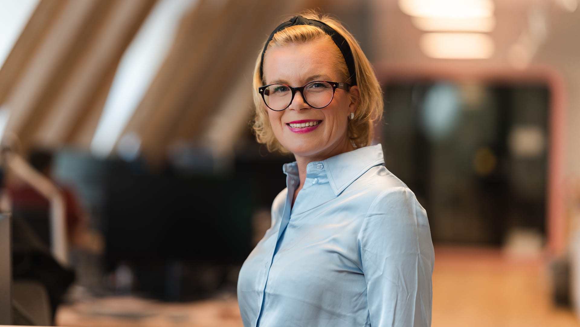 Professional woman with blonde hair wearing glasses, black headband and light blue shirt, smiling in an office setting.
