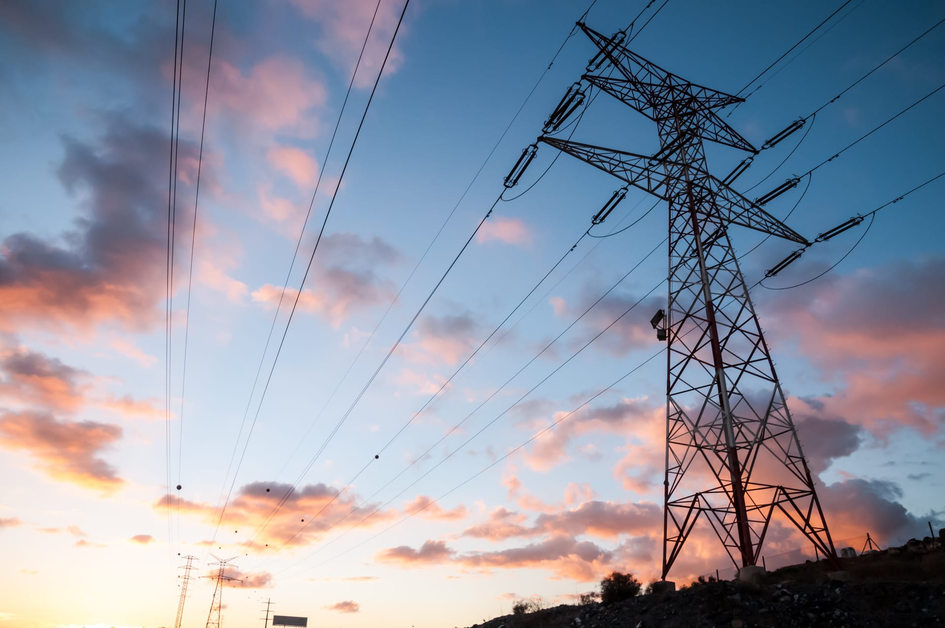 Electricity transmission tower silhouetted against a dramatic sunset sky with pink and orange clouds.