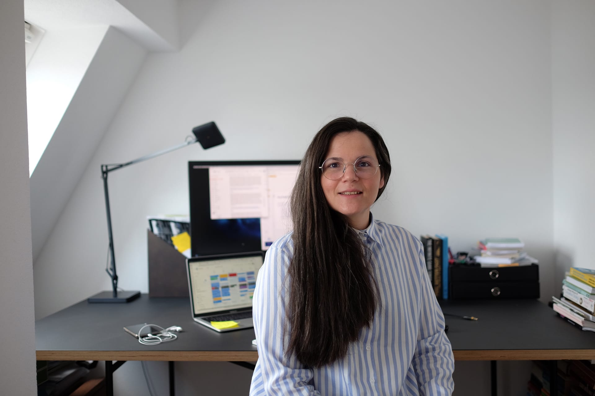 Person with long dark hair and glasses in a blue striped shirt working at a home office desk with computer monitors.