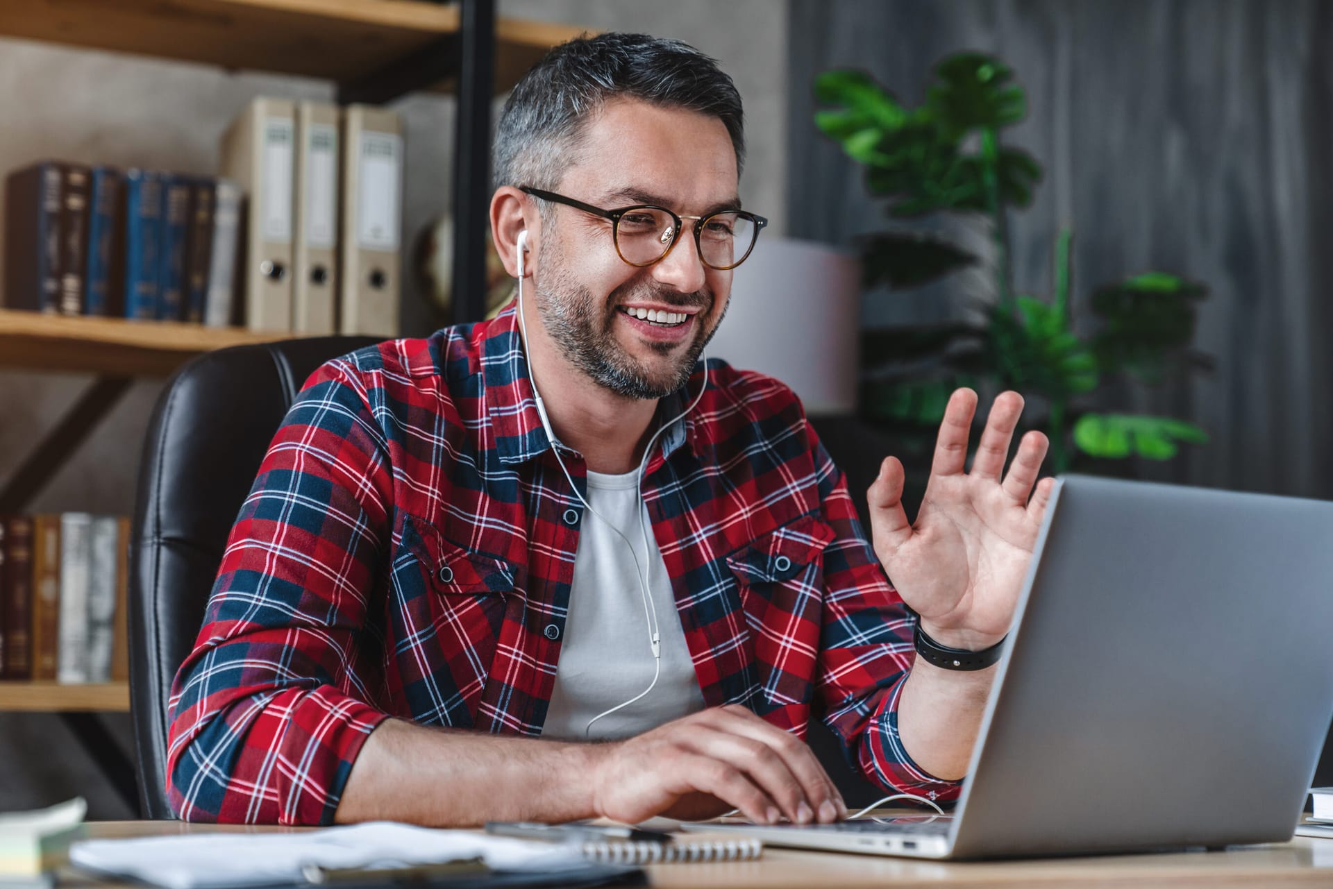 Person in red plaid shirt wearing earbuds waves during video call on laptop in home office with bookshelves and plants.