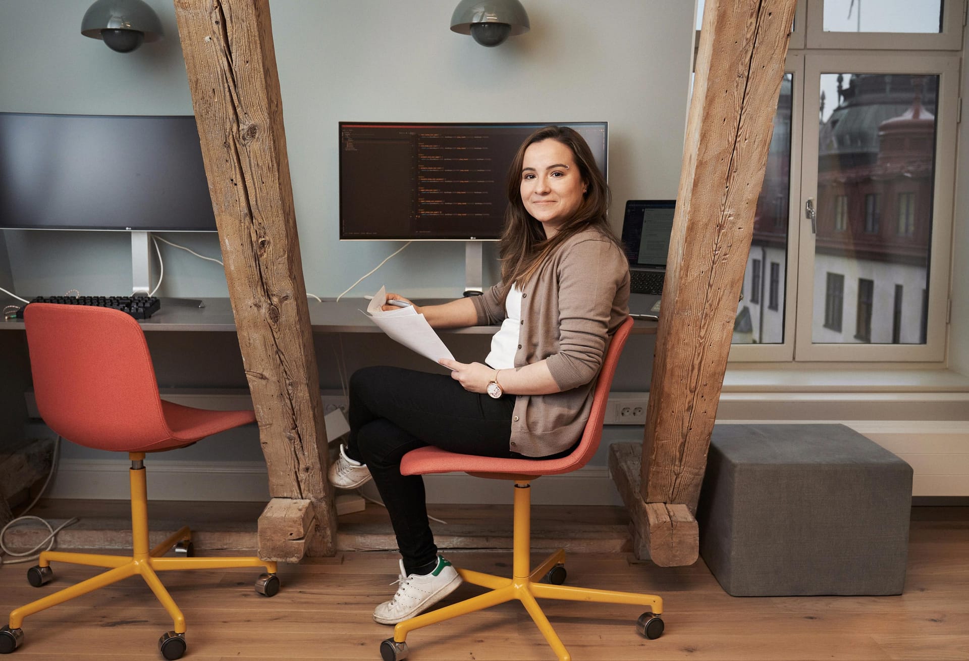 Person in orange chair working at desk with computer monitors, wooden beams, and bright window in modern office space.
