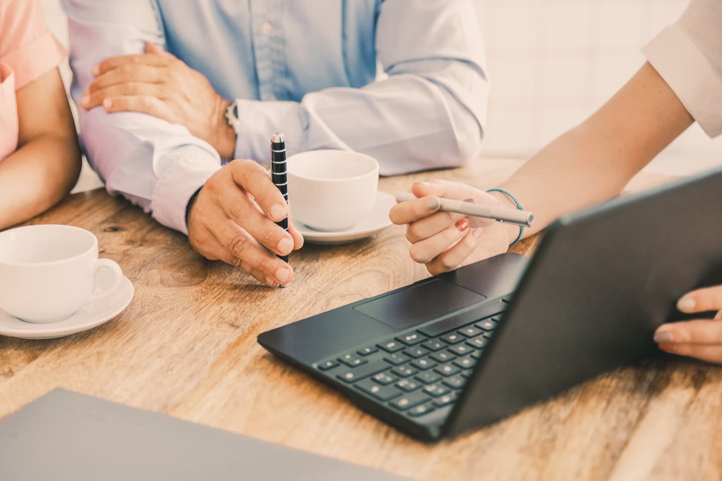 People collaborating at wooden table with laptop, coffee cups, and pens during a business meeting.
