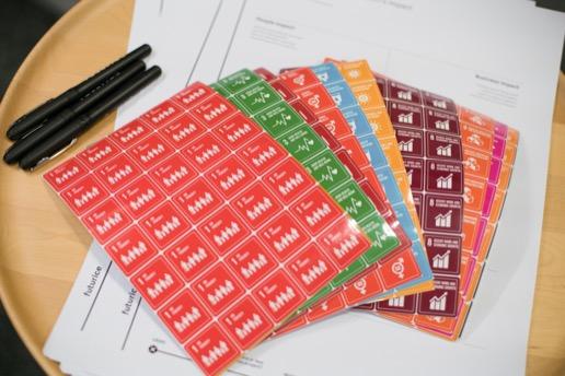 Colourful sheets of stickers for all UN SDGs, stacked and spread out, on top of impact analyzer canvas, with pencils to the side
