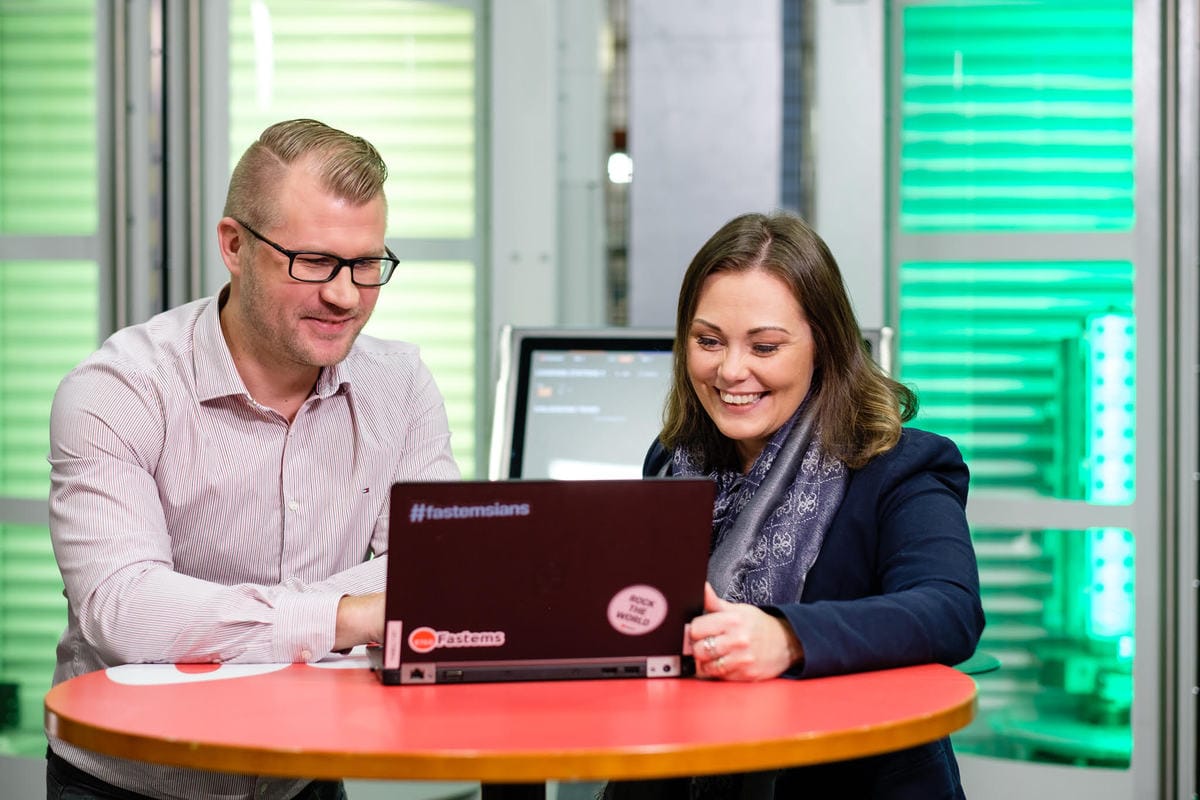 Two colleagues smiling while working together on a laptop with Fastems branding in a modern office with green lighting.