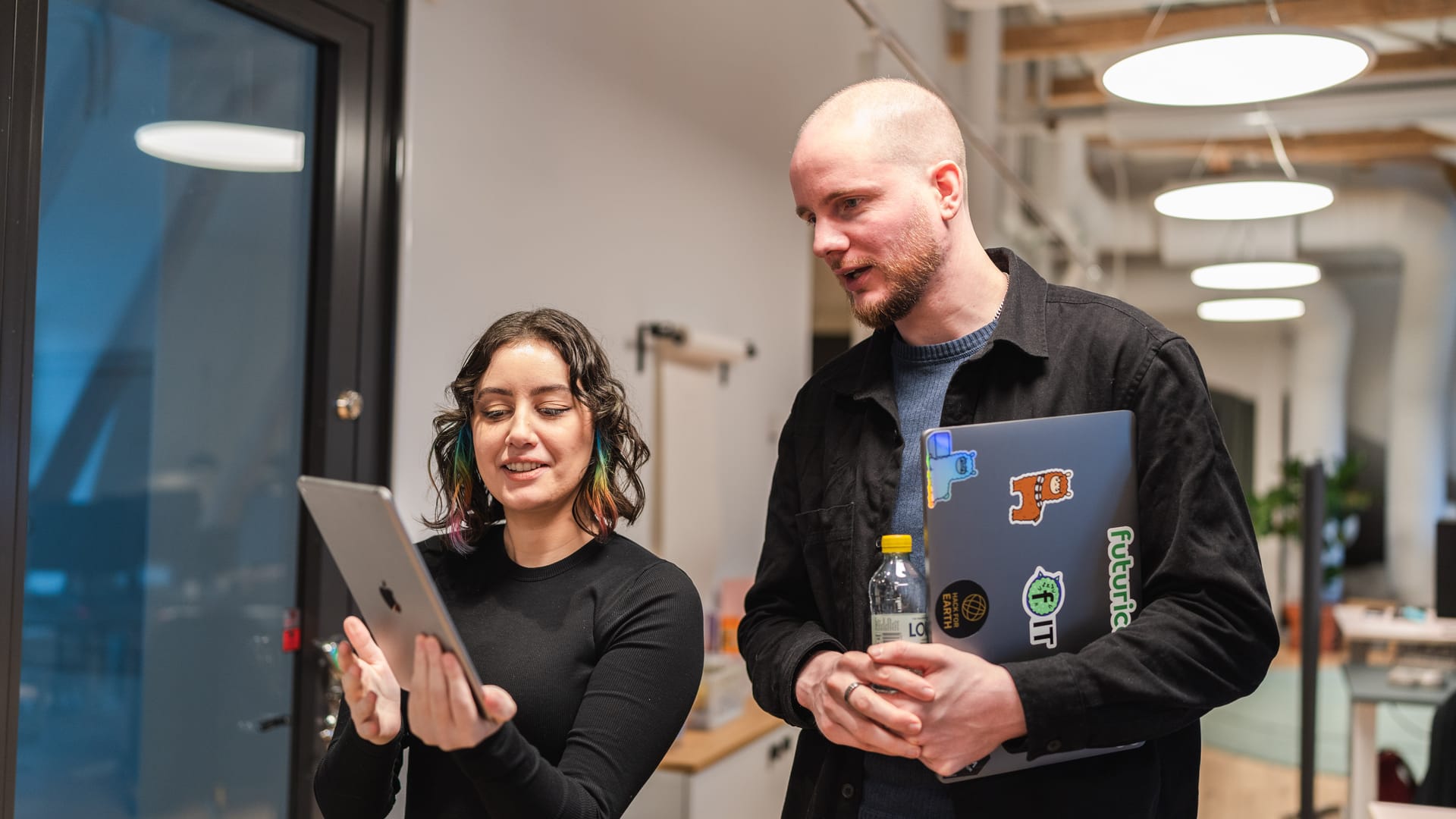 Two colleagues in a modern office looking at a tablet, one holding a laptop with colourful stickers.