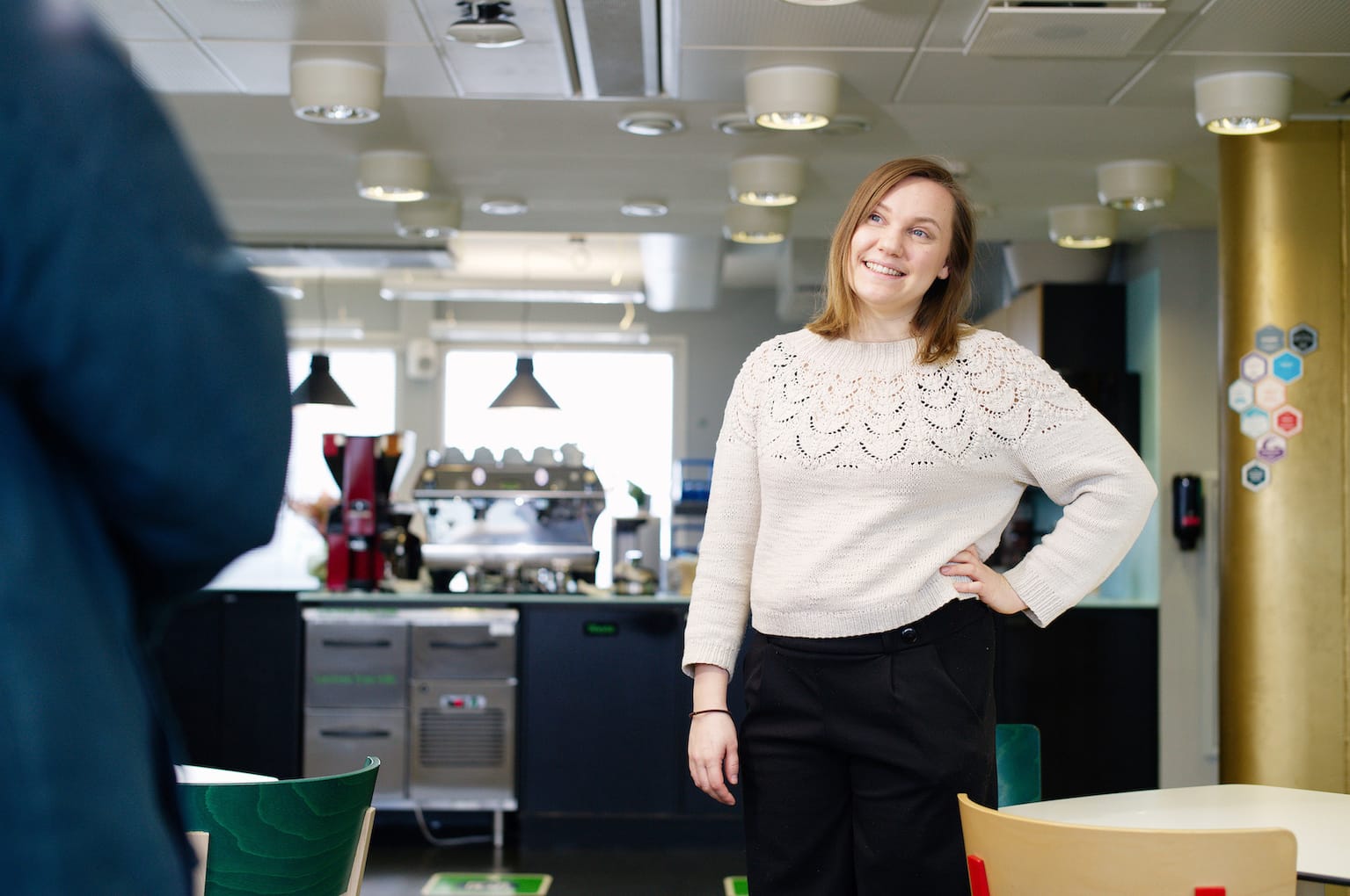 Person in white knitted jumper smiling in modern office break room with coffee area in background.