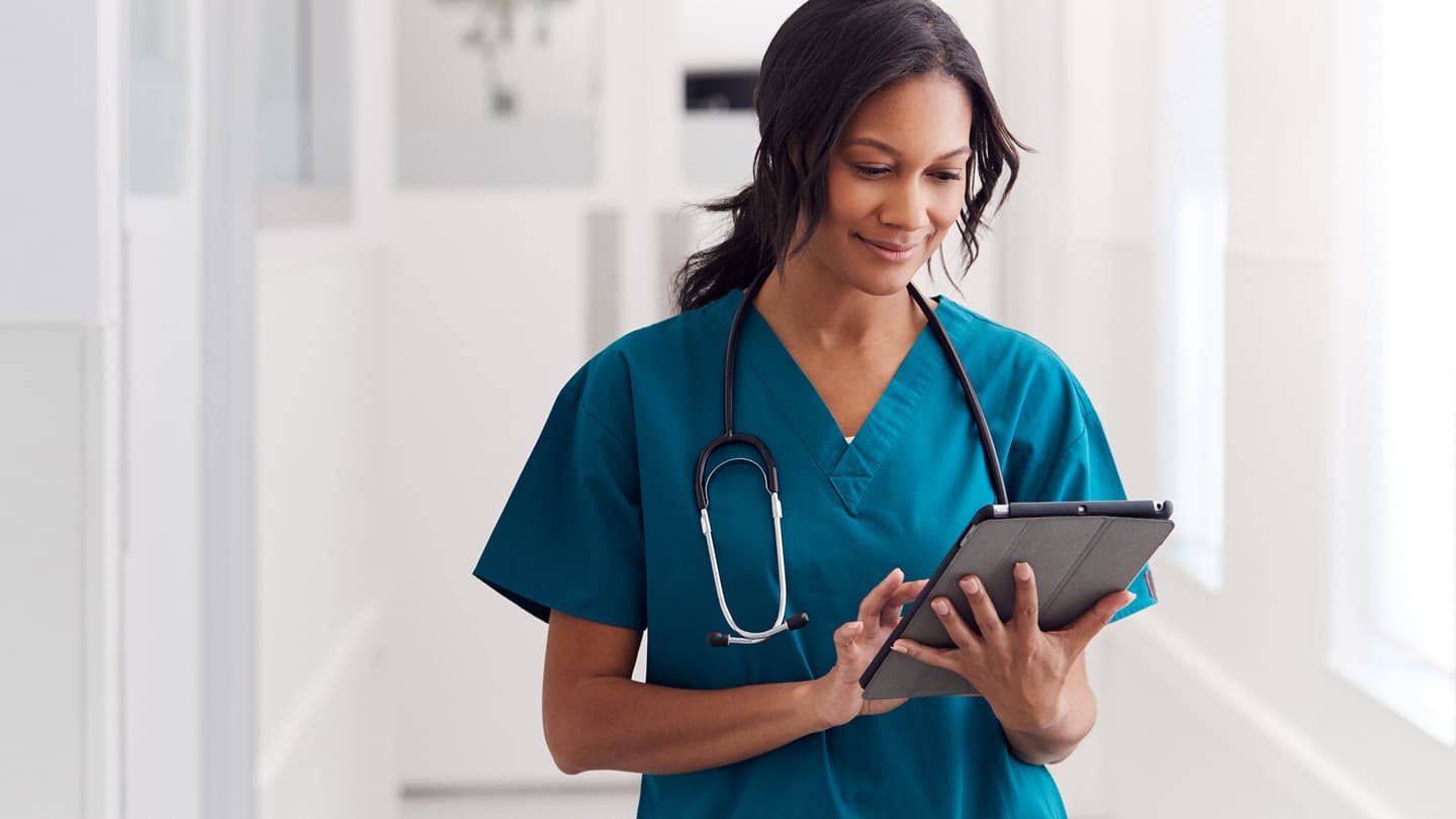 Healthcare professional in teal scrubs with stethoscope reviewing information on a tablet in a bright medical setting.