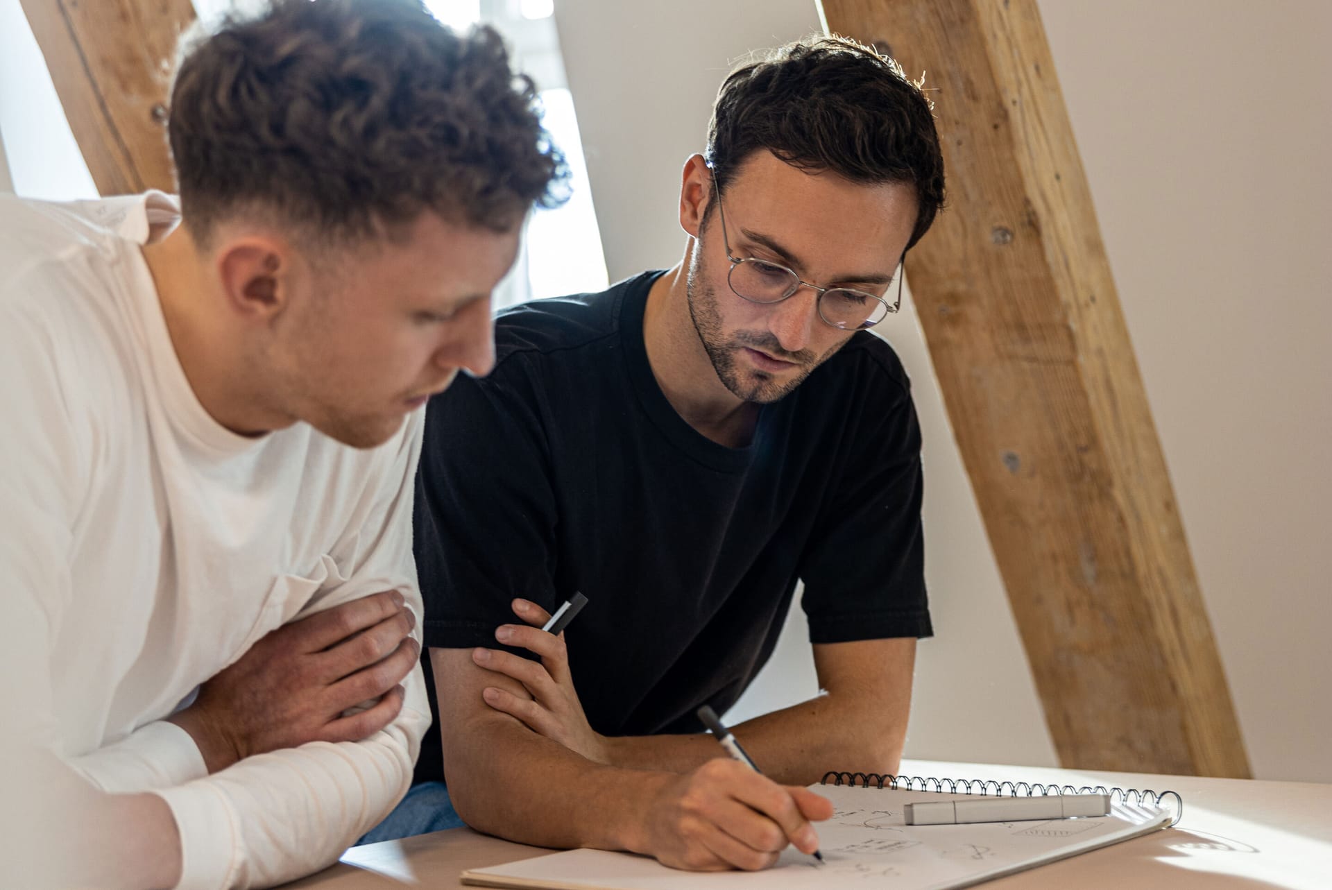 Two young men collaborating on a project, one in black t-shirt with glasses writing in a spiral notebook.