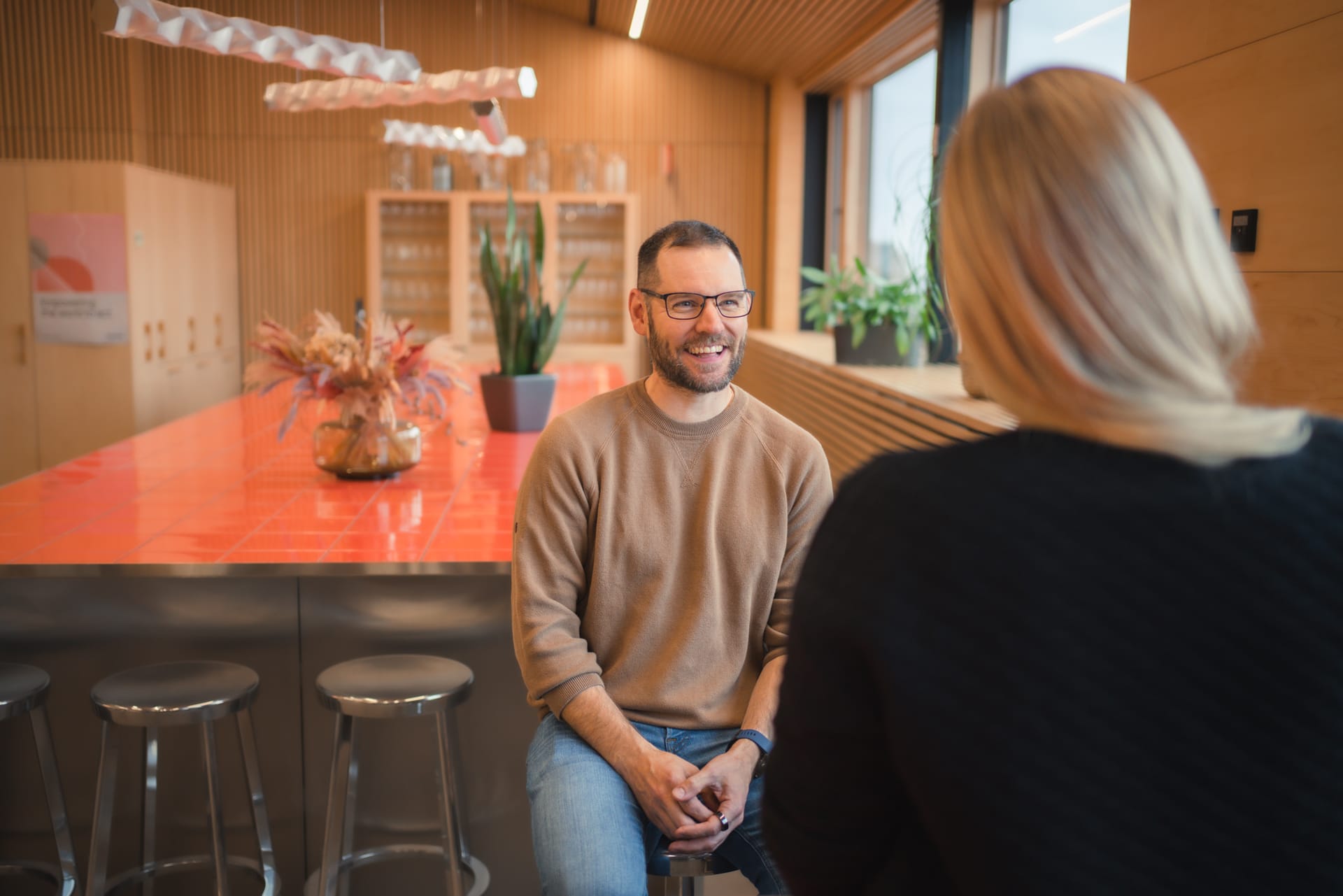 Two people conversing in a modern office space with wooden walls, orange countertop, and potted plants.