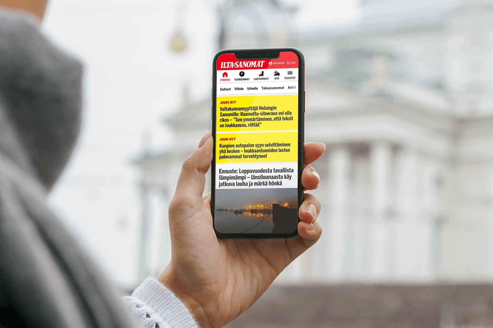 Hand holding smartphone displaying Finnish news website Ilta-Sanomat with headlines and a night harbour image.